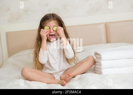 Portrait d'une fille heureuse souriante. Jolie fille de 5 ans se reposant sur le lit et faisant un masque facial avec des tranches de concombre. Concept de soins de la peau. Banque D'Images