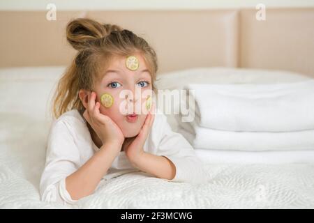 Portrait d'une fille heureuse souriante. Jolie fille de 5 ans se reposant sur le lit et faisant un masque facial avec des tranches de concombre. Concept de soins de la peau. Banque D'Images