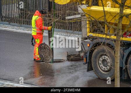 Entretien dans un trou d'homme avec un homme méconnaissable, travaillant derrière le véhicule de nettoyage d'égout sur la rue ouverte. Banque D'Images