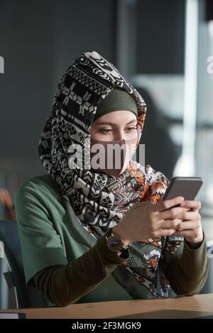 Jeune femme musulmane dans le hijab et masque de protection assis à la table et en utilisant le téléphone portable Banque D'Images