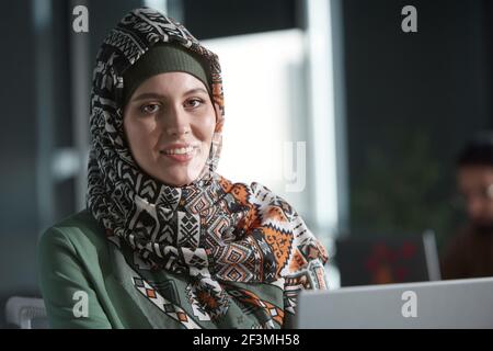 Portrait de la belle femme musulmane dans hijab souriant à la caméra assis au bureau Banque D'Images