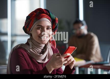 Portrait d'une jeune femme d'affaires musulmane souriant à l'appareil-photo tout en lisant un message sur son téléphone portable au bureau Banque D'Images