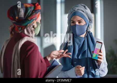 Femmes musulmanes dans des masques utilisant l'application en ligne sur téléphone mobile et travailler en équipe au bureau Banque D'Images