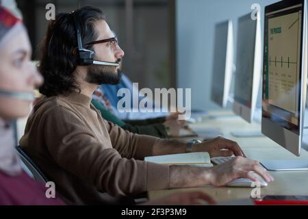 Vue latérale d'un jeune homme assis dans un casque sur le et regarder le moniteur de l'ordinateur pendant l'appel centre Banque D'Images