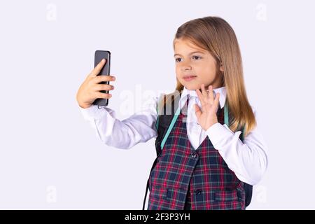 Une belle fille d'âge préscolaire en uniforme salue par la communication vidéo sur un smartphone.Formation en ligne pendant une pandémie sur fond blanc. Banque D'Images