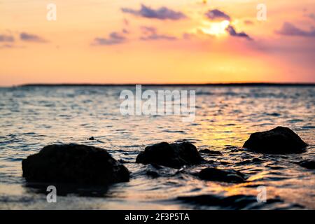 Coucher de soleil coloré à Islamorada, Florida Keys avec ciel rose, soleil derrière les nuages, reflet des vagues d'eau orange jaune, gros plan des rochers au golfe du Mexique Banque D'Images