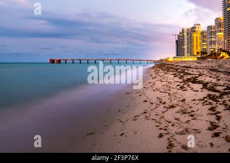 Exposition longue d'un hôtel-appartement ou d'immeubles d'appartements au crépuscule au coucher du soleil à Sunny Isles Beach de Miami, Floride par la jetée avec vagues Banque D'Images