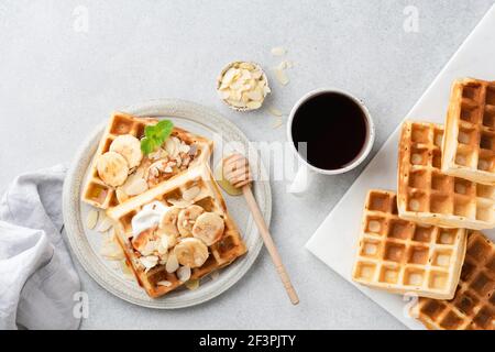 Gaufres belges et banane sucrées et café. Gaufres carrées avec banane, crème, noix d'amande et sauce caramel et tasse de café noir sur bac en béton gris Banque D'Images