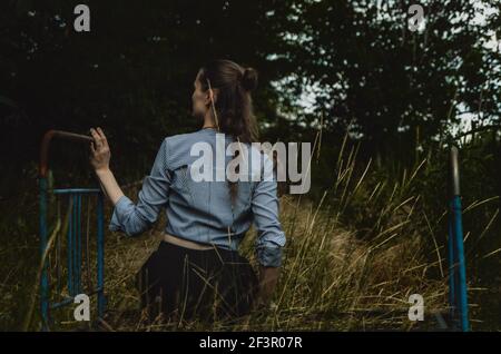 Vue arrière d'une femme avec de longs cheveux assis sur un vieux lit en métal rouillé, touchant l'herbe en croissance. Concept : mélancolie, solitude, méditation Banque D'Images