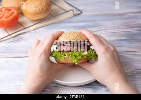 Les mains des femmes tiennent un délicieux hamburger sur fond de bois blanc avec des petits pains au sésame et de la tomate, en gros plan. Banque D'Images