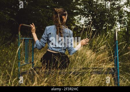 Vue arrière d'une femme avec de longs cheveux assis sur un vieux lit en métal rouillé, touchant l'herbe en croissance. Concept : mélancolie, solitude, méditation Banque D'Images