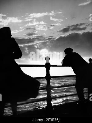 Vue d'un homme et d'une femme debout sur des rampes donnant sur la mer, silhoueté contre un soleil bas et des nuages. Plage de dates des années 1950 à 1960. Photographie de Joh Banque D'Images