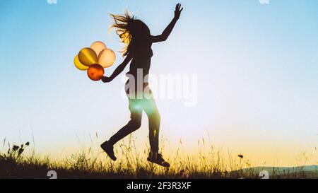 Silhouette bleu d'or d'une jeune femme sautant avec des ballons sur une colline rétroéclairé par lumière dorée de coucher de soleil, concept de liberté et de négligence Banque D'Images