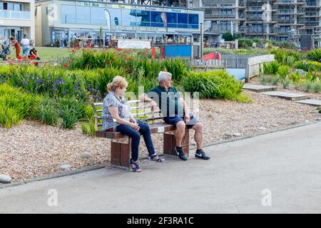 Homme et femme assis sur un banc face à la mer sur la promenade à Westward Ho!, Devon, Angleterre, Royaume-Uni, boutiques et appartements de vacances en arrière-plan Banque D'Images
