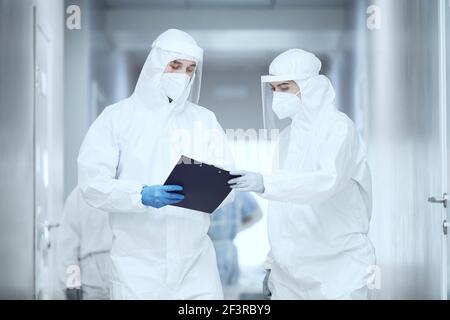 Deux médecins en uniformes de protection debout dans le couloir de l'hôpital et examen de la carte médicale ensemble Banque D'Images