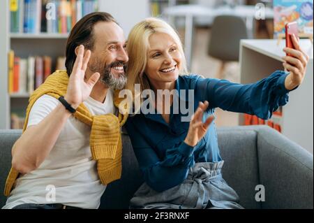 Des amis matures s'assoient sur un canapé. Homme de confiance caucasien élégant avec une barbe et une belle femme blonde sont assis sur le canapé et de discuter sur la communication vidéo à l'aide d'un smartphone, souriant Banque D'Images