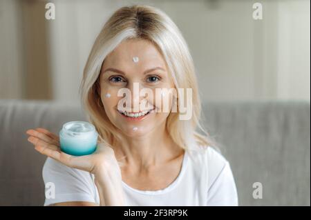 La femme adulte blonde caucasienne sourit et regarde la caméra. Femme adulte incroyable tient une crème dans la main, crème hydratante du visage dans son visage, applique des gouttes de la crème sur son visage, soins de peau Banque D'Images