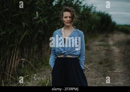 Portrait d'une jeune femme caucasienne à expression neutre, près des roseaux sur un chemin, moody Vibe Banque D'Images