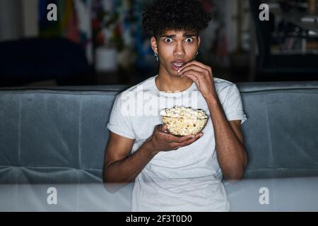 Un jeune homme effrayé avec les yeux ouverts, portant un pyjama et mangeant du pop-corn tout en regardant un film d'horreur, assis sur un canapé confortable à la maison Banque D'Images