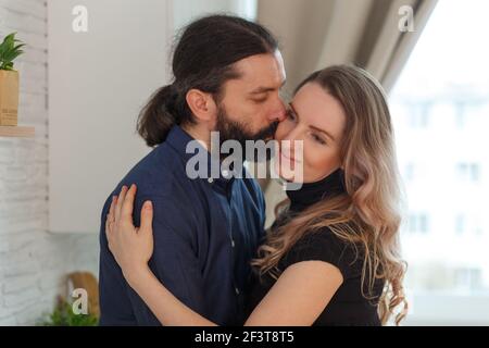 Homme embrassant partenaire enceinte dans la cuisine. Concept de grossesse familiale adulte. Les futurs parents dans la tenue à la maison embrassent debout dans la cuisine, regardant e Banque D'Images