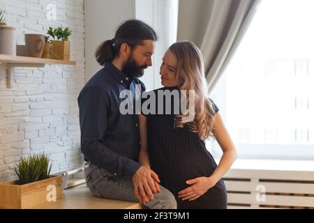 Homme embrassant partenaire enceinte dans la cuisine. Concept de grossesse familiale adulte. Les futurs parents dans la tenue à la maison embrassent debout dans la cuisine, regardant e Banque D'Images