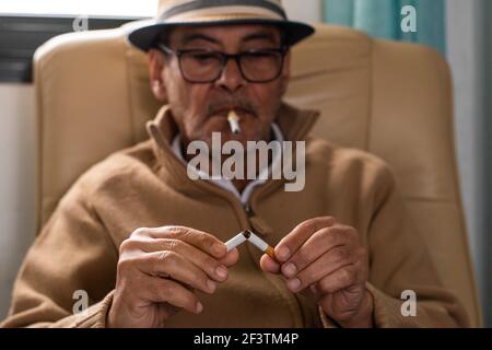 Homme caucasien âgé portant un chapeau et des lunettes noires, tenant dans sa bouche une cigarette en plastique pour arrêter de fumer, et dans ses mains un cigare qu'il a Banque D'Images