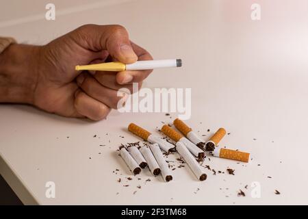 Main d'un homme tenant une cigarette en plastique pour arrêter de fumer, à côté d'une pile de cigarettes cassées comme signe de rejet du tabac sur une table blanche. Banque D'Images