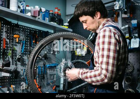 Expertise technique prenant soin de la boutique de vélos. Beau jeune mécanicien fixant la roue de cycle dans l'atelier. Beau réparateur en vêtements de travail servant la montagne Banque D'Images