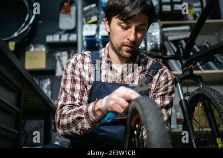 Expertise technique prenant soin de la boutique de vélos. Beau jeune mécanicien fixant la roue de cycle dans l'atelier. Beau réparateur en vêtements de travail servant la montagne Banque D'Images