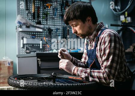 Expertise technique prenant soin de la boutique de vélos. Beau jeune mécanicien fixant la roue de cycle dans l'atelier. Beau réparateur en vêtements de travail servant la montagne Banque D'Images