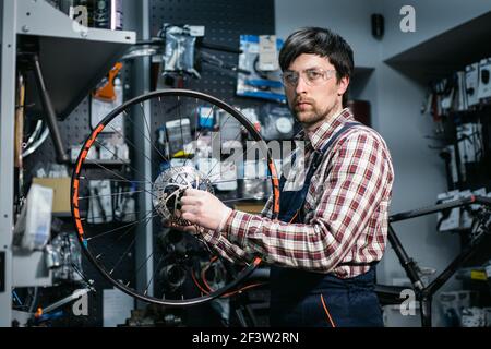 Expertise technique prenant soin de la boutique de vélos. Beau jeune mécanicien fixant la roue de cycle dans l'atelier. Beau réparateur en vêtements de travail servant la montagne Banque D'Images