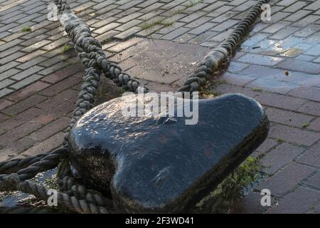 Un bollard mouillage humide peint en noir, avec trois cordes d'amarrage tressées épaisses de navires amarrés attachées à lui, monté sur un quai recouvert de gris et Banque D'Images