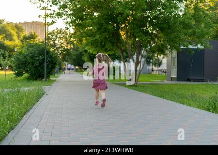 une petite fille dans une robe à pois court gaiement dans un saut le long de la ruelle du parc un jour d'été, vue arrière Banque D'Images