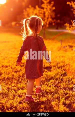 Une petite fille caucasienne dans une robe marche dans le parc dans les rayons du soleil couchant Banque D'Images