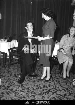 1957, historique, à l'intérieur d'une salle de réception dans un hôtel, une jeune femme attrayante portant un plateau avec une boisson sur lui pour un homme dans une veste de dîner, peut-être le chef de groupe, à l'amusement d'une femme assise à proximité, Victoria Hotel, Leeds, Angleterre, Royaume-Uni. Banque D'Images