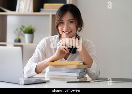 Portrait d'une femme asiatique souriante assise à une table dans la pièce, regardant l'appareil photo, une femme excitée posant, travaillant à l'ordinateur, faisant des devoirs Banque D'Images