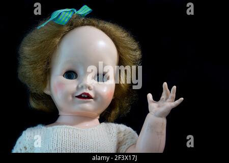 Vieille poupée antique en plastique dur avec des cheveux blonds et des yeux de regard et en agitant un bras contre un fond noir. Banque D'Images