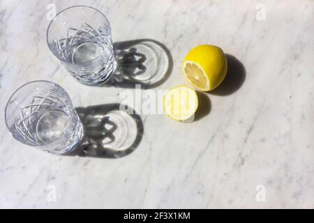 Deux verres d'eau et un citron dans la lumière du soleil avec de longues ombres sur un fond texturé de marbre gris et blanc vue de dessus vers le bas et mise au point intentionnelle. Banque D'Images