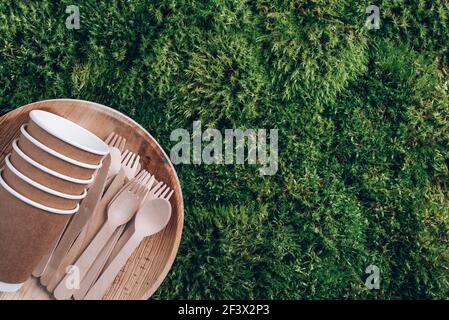 Vaisselle jetable en matériaux naturels. Assiettes en bambou, cuillère en bois, fourchette, couteau, gobelets en papier sur fond de mousse verte. Écologique et durable Banque D'Images