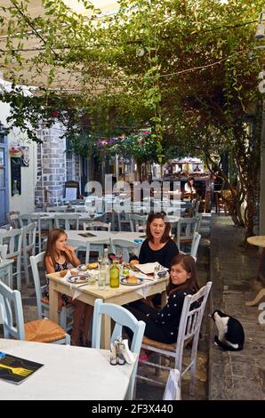 Une famille de touristes mange le déjeuner dans un restaurant à Ermoupoli, Syros, Cyclades, Grèce, comme un chat errant regarde dessus Banque D'Images