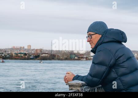 Portrait en gros plan d'un homme d'âge moyen en lunettes debout sur le remblai de la rivière et regardant au loin. Un homme dans une veste et un chapeau chauds. Banque D'Images