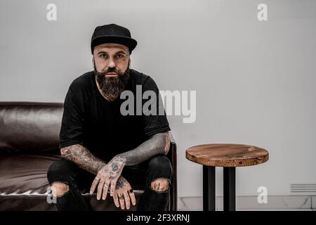Un jeune artiste de tatouage masculin avec une barbe a l'air sérieux assis sur un canapé dans un lieu d'atelier. Banque D'Images