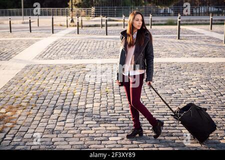 jeune femme avec valise à roulettes marchant dans une rue pavée, concept de voyage et de style de vie urbain, espace de copie pour le texte Banque D'Images