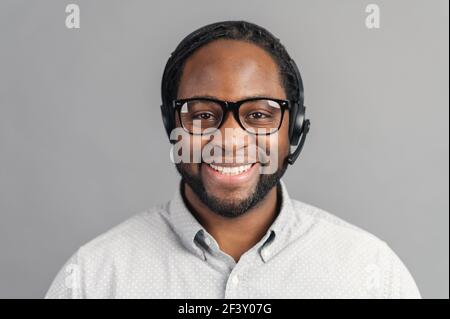 Jeune travailleur de bureau afro-américain souriant en lunettes et casque regardant la caméra et souriant, homme noir positif travaillant dans le service à la clientèle, isolé sur le gris Banque D'Images