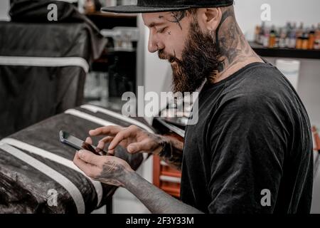 Jeune artiste de tatouage masculin avec la barbe vérifiant son téléphone assis sur le canapé dans l'atelier. Banque D'Images