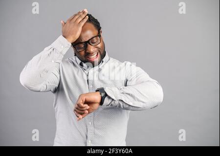 Un homme d'affaires afro-américain surpris portant une chemise élégante et décontractée et des lunettes est tard, regarde la montre de main et tient la tête frustré, un employé de bureau a oublié de se rencontrer, isolé sur le gris Banque D'Images