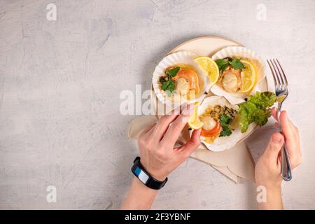 Les mains des femmes tiennent la fourchette et écrasez le citron sur les coquilles Saint-Jacques. Noix de Saint-Jacques cuites au caviar dans une assiette blanche sur fond de pierre blanche.dîner avec fruits de mer. Banque D'Images