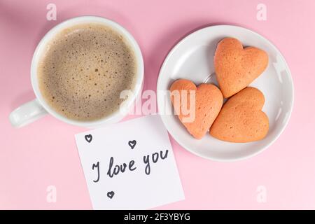 Note avec les mots que je vous aime avec des biscuits en forme de coeur et de tasse de café sur couleur jaune pastel. Petit déjeuner le matin ou le jour de la Saint-Valentin Banque D'Images