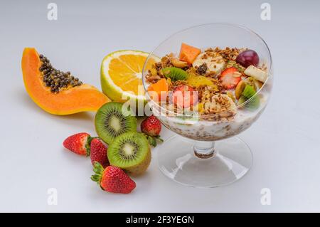 Yaourt avec granola et fruits comme fraise, raisin, banane, papaye, orange, kiwi dans un bol en verre sur fond blanc. Une alimentation saine. Banque D'Images