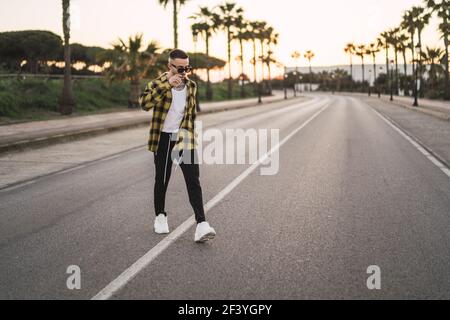 Un cliché peu profond d'un beau homme espagnol de race blanche avec des tatouages et des lunettes de soleil traversant la rue pendant le coucher du soleil Banque D'Images